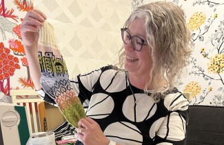 Villasukkakilpailun voittaja Taina Miettinen esittelee Strategiasukkaa iloisena. Edessä pöydällä on useampia lankarullia ja taustalla näkyy hyvinvointialueen strategiakuva.