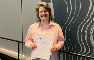 Henkilö hymyilee kameralle ja pitää molemmin käsin kiinni A4 kokoisesta paperista, jossa lukee palkinnon nimi.