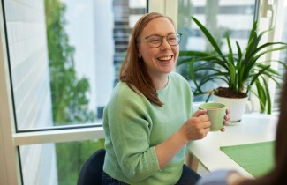 Nuori nainen hymyilee pöydän ääressä istuen ja pitelee kädessään mukia.