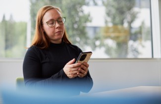 Kuvassa aikuinen naisoletettu katsoo kännykkäänsä. Naisella on pitkät, punertavat hiukset ja musta poolupaita. Nainen istuu istuu tuolilla pöydän ääressä. Takana olevasta ikkunasta näkyy metsämaisema.