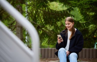 Nuori nainen katsoo hymyillen älypuhelinta. Taustalla on vihreitä puita.
