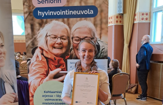 Nainen hymyilee kunniakirja kädessä seniorin hyvinvointineuvolan roll-upin edessä.