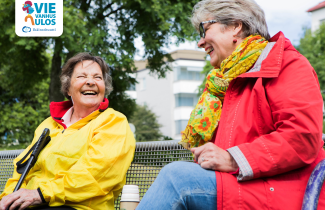 Kaksi iloista ikäihmistä istuu ulkona penkillä.  Vie vanhus ulos -kampanjan kuva.