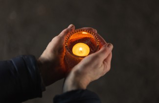 Kaksi kättä pitää tuikkukippoa, jossa palaa lämmin tuikku.