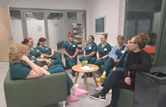 Yhdeksän hoitajan työasuun pukeutunutta osallistujaa istuu pyöreän pöydän ääressä sohvilla. 