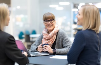 Kolme henkilöä istuu työpöydän ääerssä. Yksi heistä katsoo kameraan ja hymyilee.