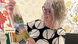 Villasukkakilpailun voittaja Taina Miettinen esittelee Strategiasukkaa iloisena. Edessä pöydällä on useampia lankarullia ja taustalla näkyy hyvinvointialueen strategiakuva.