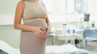 Raskaana oleva henkilö pitelee käsistä muodostettua sydäntä vatsansa päällä, nainen on kuvattu hartioista alaspäin.