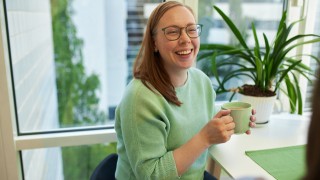 Nuori nainen hymyilee pöydän ääressä istuen ja pitelee kädessään mukia.