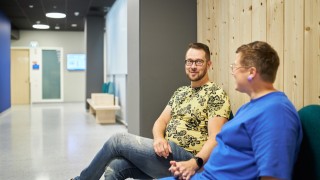 Kaksi ihmistä istuu vieretysten penkillä katsoen toisiaan kohden ja pidellen kädestä kiinni. 