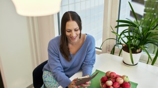 Henkilö puhelimella pöydän ääressä, omenakulho ja viherkasvi taustalla.