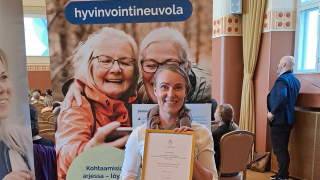 Nainen hymyilee kunniakirja kädessä seniorin hyvinvointineuvolan roll-upin edessä.