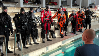 Vesisukeltajakoulutettavat sukelluspuvuissa uima-altaan reunalla