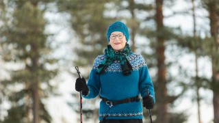 Iäkäs nainen sauvakävelee villapaita päällä ja taustalla on puita