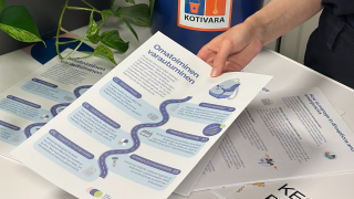 Henkilö ojentaa esitettä, jossa käsitellään omatoimista varautumista. Taustalla ämpäri, jonka kyljessä tarra, jossa lukee Kotivara