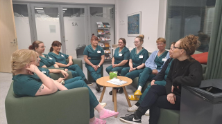 Yhdeksän hoitajan työasuun pukeutunutta osallistujaa istuu pyöreän pöydän ääressä sohvilla. 