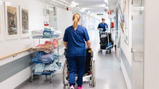 Hoitaja kuljettaa pyörätuolissa istuvaa potilasta terveyskeskuksen käytävällä. Käytävän varrella on siivouskärry ja siivooja.